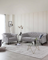 Elegant living room featuring a grey tufted sofa with a curved back, complemented by a matching tufted accent armchair. A round coffee table with a glass top and unique gold-coloured metal base is centred on a textured light grey area rug.