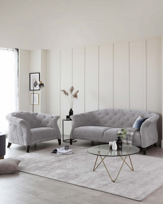 Elegant living room featuring a grey tufted sofa with a curved back, complemented by a matching tufted accent armchair. A round coffee table with a glass top and unique gold-coloured metal base is centred on a textured light grey area rug.