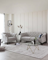 Elegant living room set featuring a large, tufted-back, grey fabric sofa, accompanied by a matching armchair with a similar design. In front of the sofa is a round, glass-top coffee table with a gold-finished geometric metal base. The set is arranged on a textured grey area rug, accentuating the modern yet sophisticated atmosphere of the space.