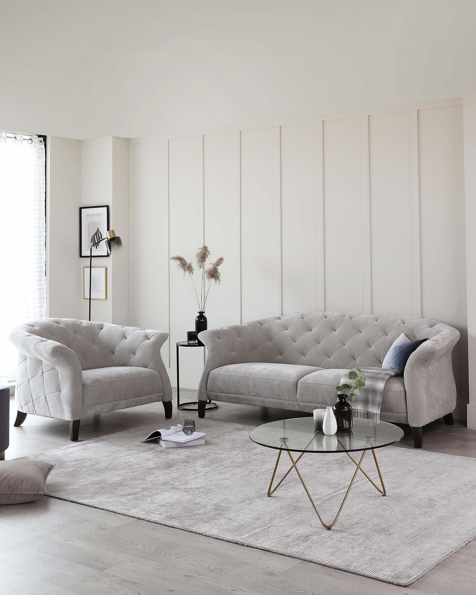 Elegant living room set featuring a large, tufted-back, grey fabric sofa, accompanied by a matching armchair with a similar design. In front of the sofa is a round, glass-top coffee table with a gold-finished geometric metal base. The set is arranged on a textured grey area rug, accentuating the modern yet sophisticated atmosphere of the space.