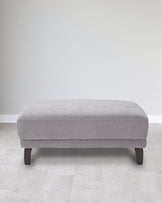 Grey upholstered ottoman with curved edges and dark wooden legs, set against a light, minimalist background.