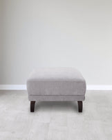 A modern, square, light gray upholstered ottoman with dark wood legs, set against a minimalist background.