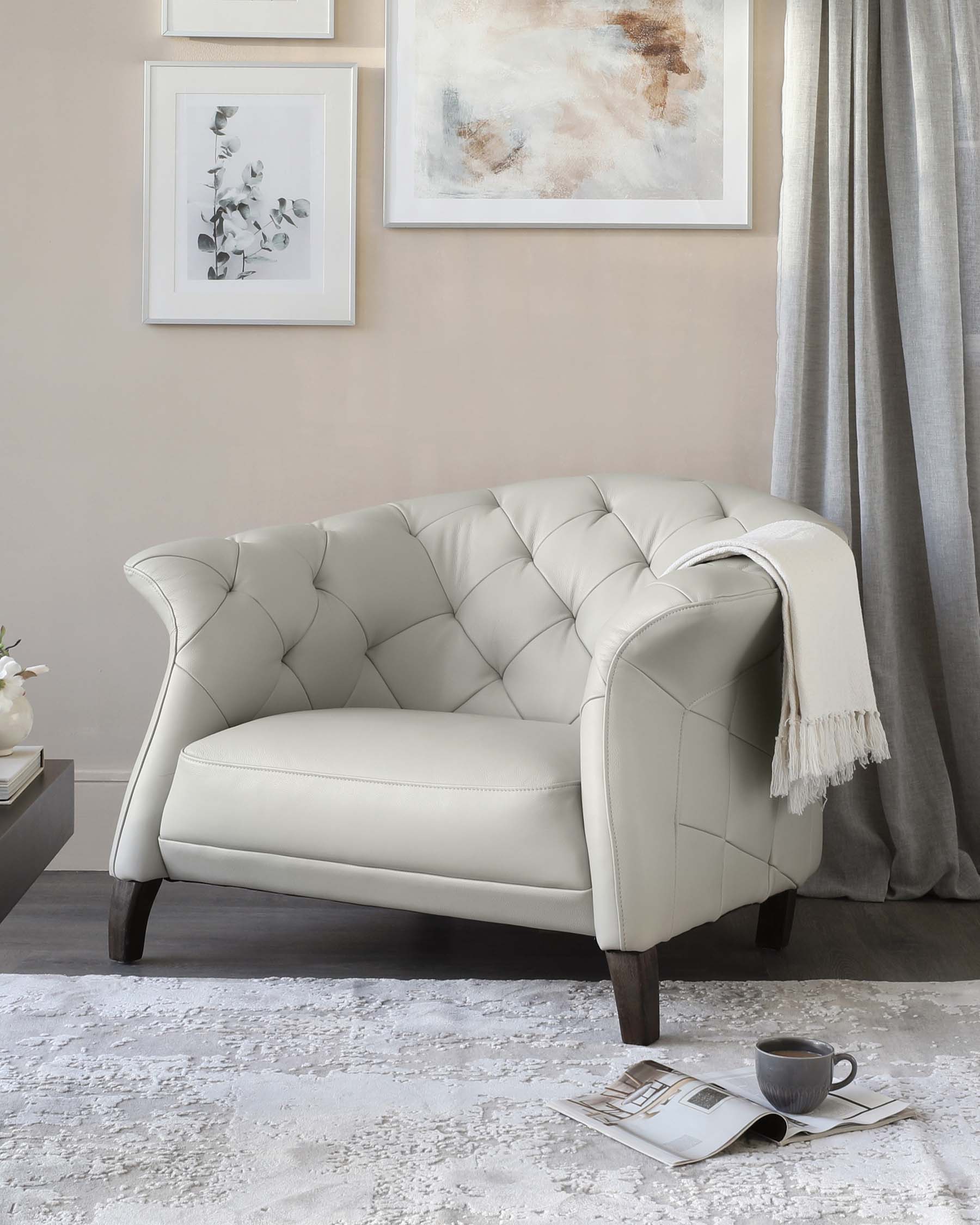 Elegant tufted cream loveseat with a curved backrest and rolled arms, featuring diamond button detailing and dark wooden legs, accessorized with a light grey fringe throw blanket on one arm, set against a light neutral-toned room with wall art, a grey curtain, and a white fluffy area rug. A small side table with a white vase and books, along with a black coffee cup and a magazine on the floor, complement the scene.