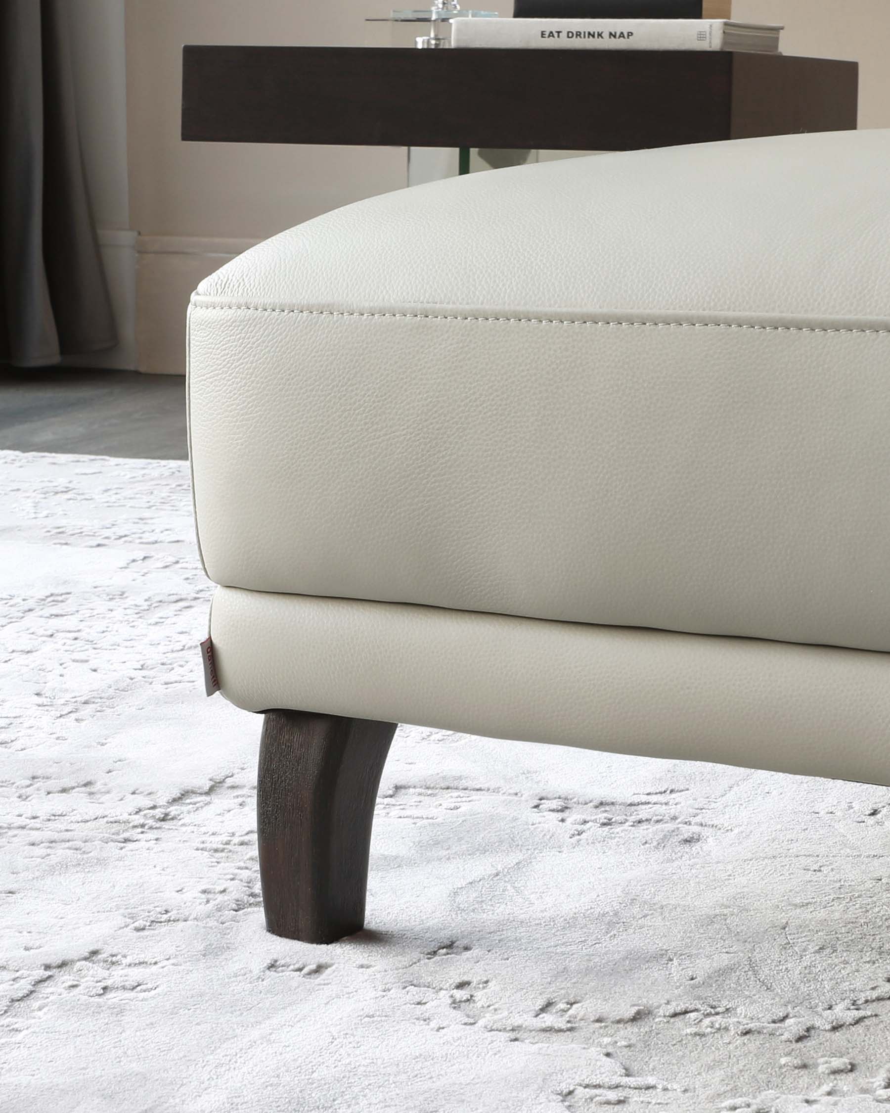 Close-up of a contemporary sofa with cream textured upholstery and elegant dark wooden tapered legs, set against a plush grey rug. A sleek dark console table looms in the background with a minimalist display.