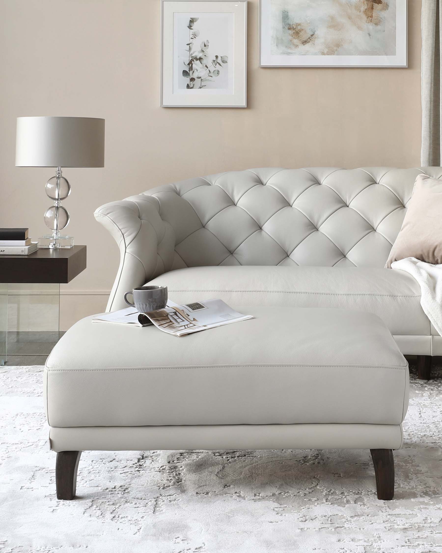 Elegant contemporary living room set featuring a cream-colored tufted chesterfield sofa with a matching ottoman, dark wooden legs, accented with a soft beige cushion and a light throw. A sleek side table with a modern lamp and framed artwork adds to the refined ambiance.