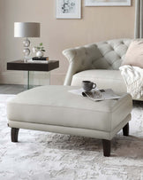 Elegant living room featuring a tufted, cream-colored leather sofa with a plush throw pillow and fringed blanket, a modern leather ottoman with dark wooden legs holding a tray with a teacup and reading materials, and a sleek black side table with a glass top supporting a decorative lamp, books, and a small plant. The room is accented with a soft, textured area rug and a simple framed artwork on the wall.