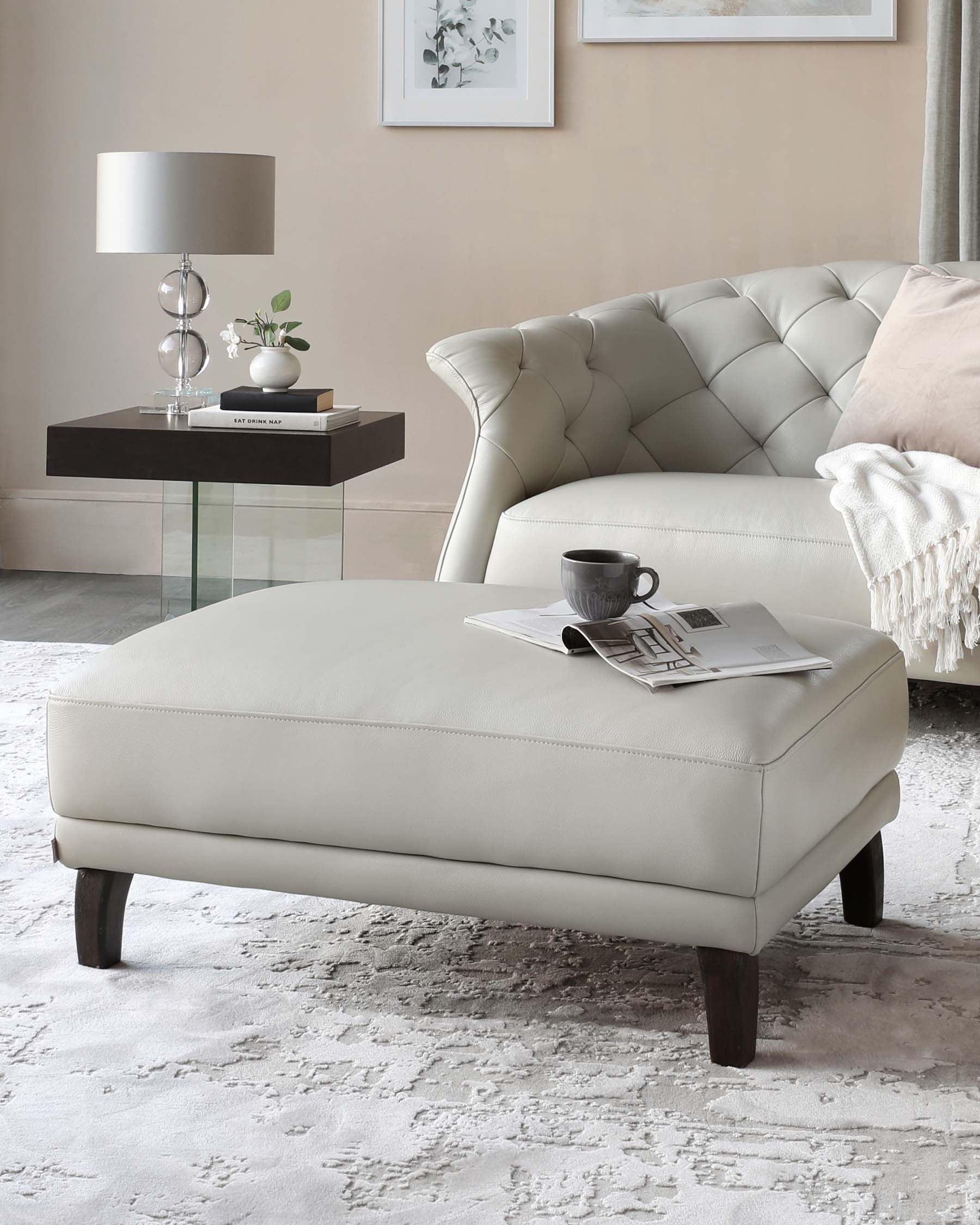 Elegant living room featuring a tufted, cream-colored leather sofa with a plush throw pillow and fringed blanket, a modern leather ottoman with dark wooden legs holding a tray with a teacup and reading materials, and a sleek black side table with a glass top supporting a decorative lamp, books, and a small plant. The room is accented with a soft, textured area rug and a simple framed artwork on the wall.