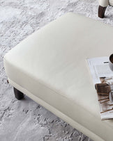 A contemporary cream-colored leather ottoman with a smooth finish and visible stitching details on the sides. It has dark, short wooden legs. On top of the ottoman lies an open magazine, with a coffee cup placed on it, suggesting a relaxed living space. The ottoman is positioned on a textured grey area rug.