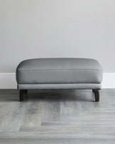 Modern grey upholstered ottoman with a rectangular cushioned top and four dark wooden legs, set against a neutral wall and light grey wooden flooring.