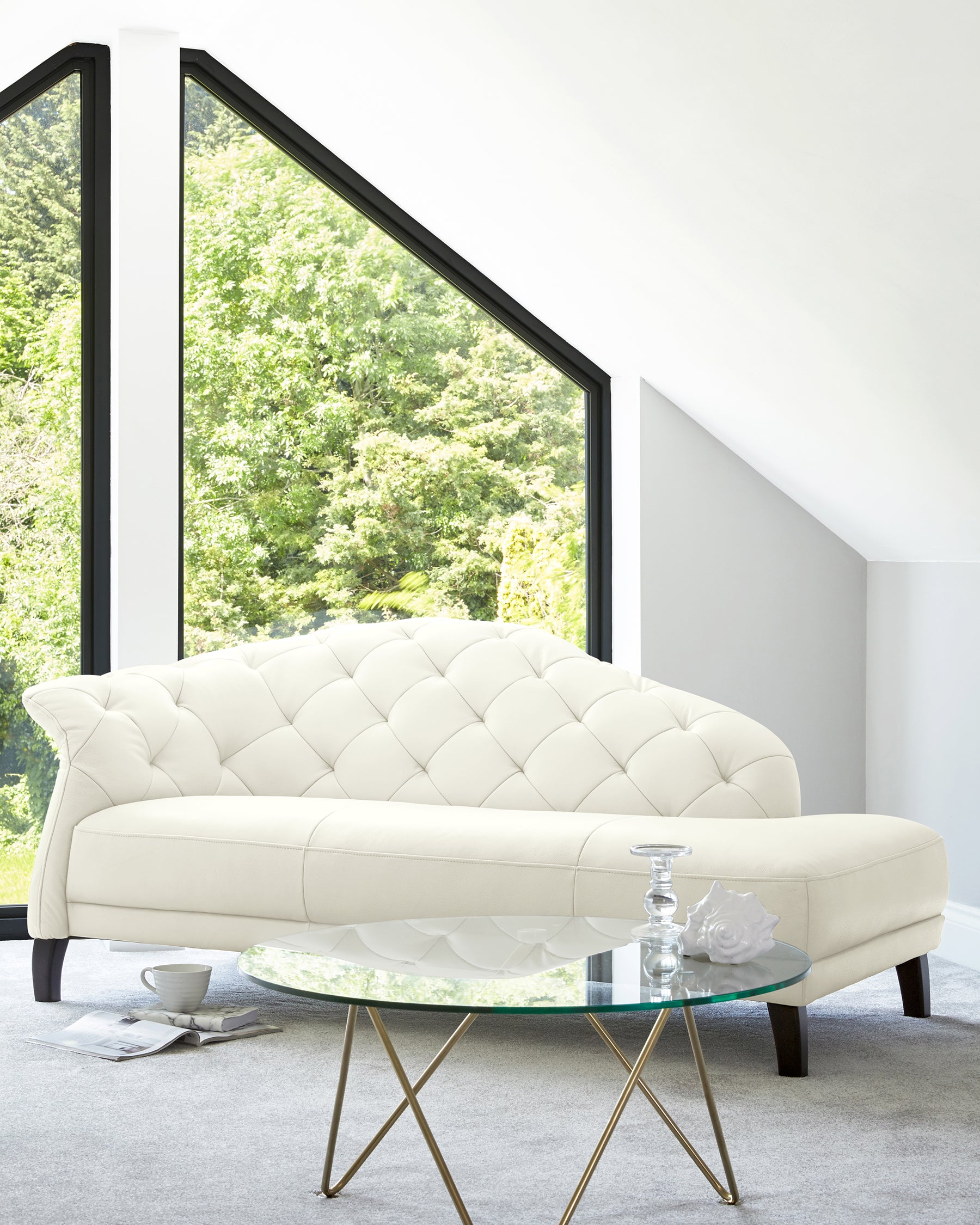 Elegant white tufted sofa paired with a round glass coffee table featuring golden legs, set in a bright room with large windows.