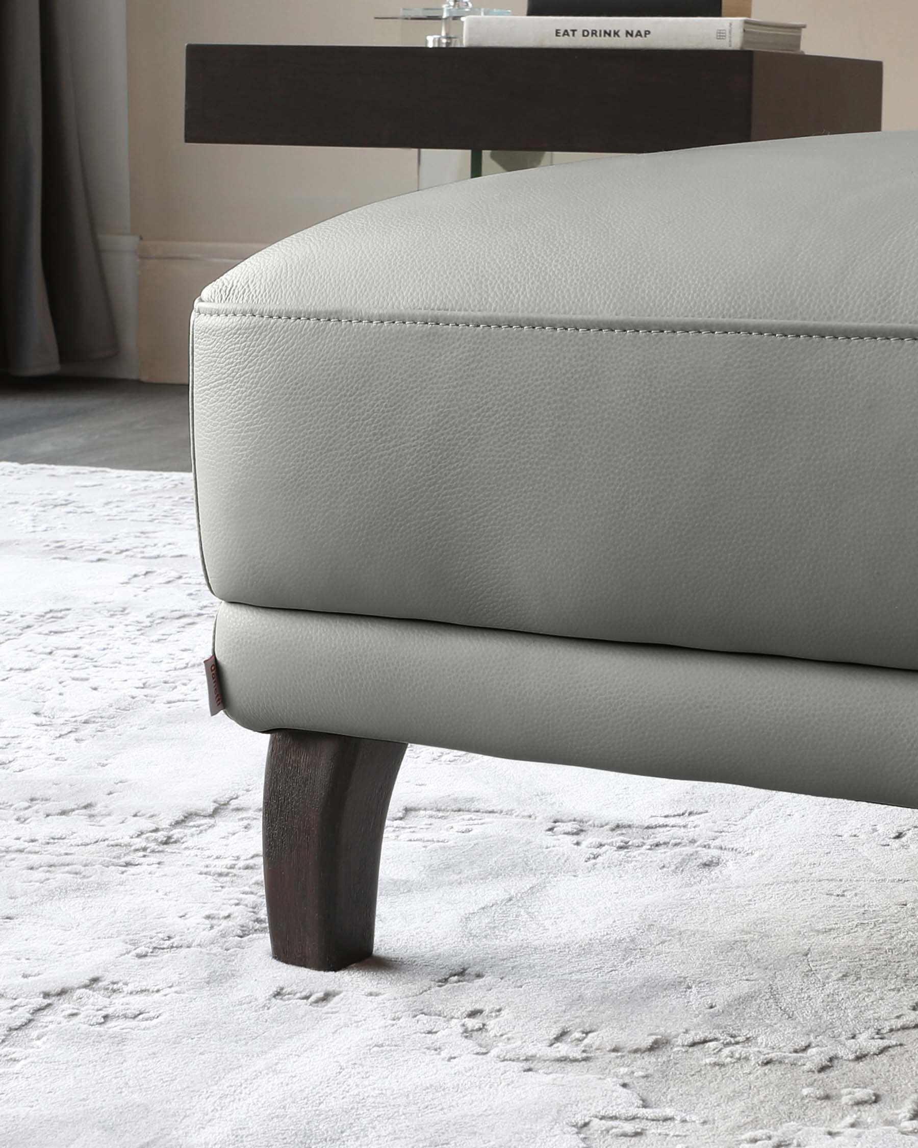 Modern light grey upholstered ottoman with visible stitching details and dark wooden tapered legs, shown in a contemporary room setting with a shaggy white rug, partially visible dark wood side console with books, set against a neutral wall and window drapes.