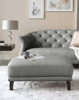 Elegant grey tufted chesterfield sofa with matching ottoman, featuring a quilted design and dark wooden legs, accessorized with a soft throw blanket, decorative cushion, and a coffee cup resting on a tray with reading materials.