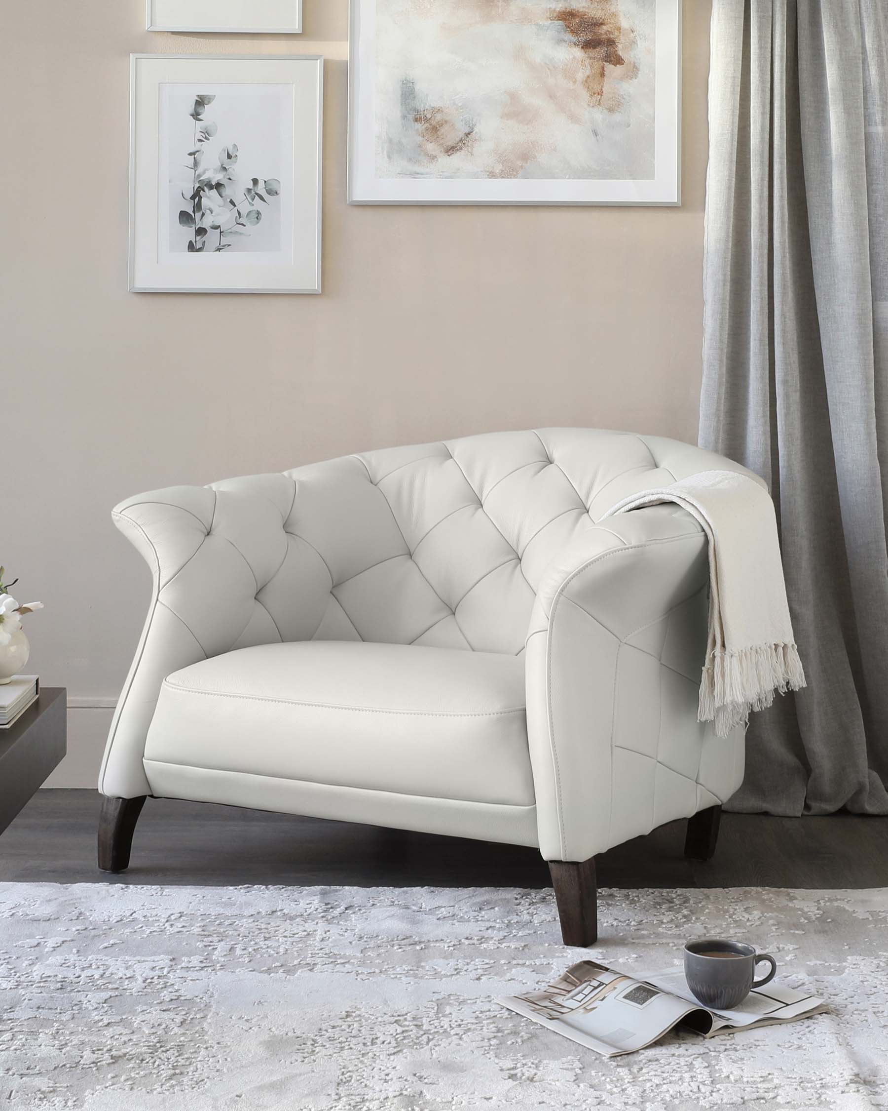 Elegant ivory-coloured tufted loveseat with curved backrest and arms, featuring button detailing and sitting on dark wooden legs, accented by a soft white throw blanket over one arm. Positioned on a textured white area rug with a casual arrangement of lifestyle magazines and a grey ceramic mug beside it, providing a cosy and inviting atmosphere.