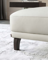 Ivory leather-upholstered ottoman with dark wooden tapered legs and fine stitch detailing, placed on a textured light grey rug, with a dark wood sideboard and decorative books in the background.