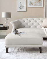 An elegant, cream-colored tufted chesterfield sofa with matching ottoman, featuring classic rolled arms and dark wooden legs. The sofa is complemented by a contemporary lamp on a dark bedside table, and a pristine area rug underneath. The scene is accented by soft decorative pillows, a casual throw, and a touch of sophistication with an art frame and magazine on the ottoman.