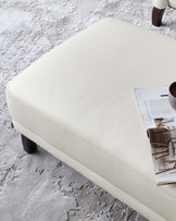 Elegant ivory leather ottoman with a smooth finish and slightly raised edges, featuring dark wooden block legs, complemented with a soft grey textured rug underneath. Includes a magazine for added ambiance.