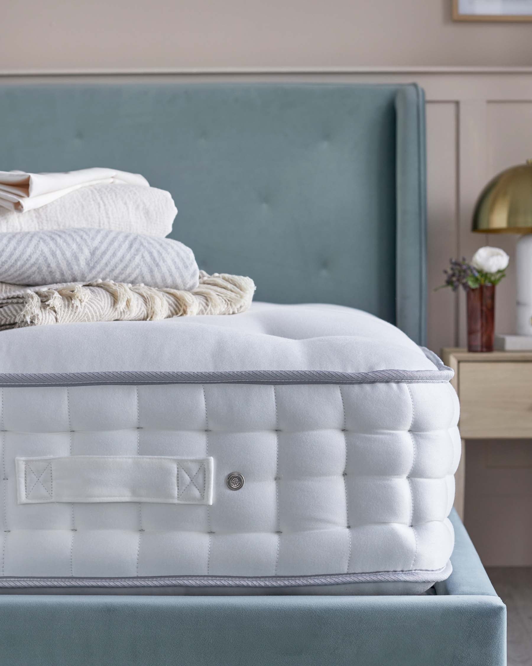 Elegant bedroom scene focusing on a luxurious upholstered bed frame in a soft teal colour with a padded, tufted headboard that adds a sense of comfort and sophistication. A crisp white mattress is prominently featured, topped with folded white linens and a cosy beige throw blanket, highlighting the bedding products available. In the background, a wooden bedside table with a golden lamp and small decorative items can be seen, creating an inviting and harmonious sleeping environment.