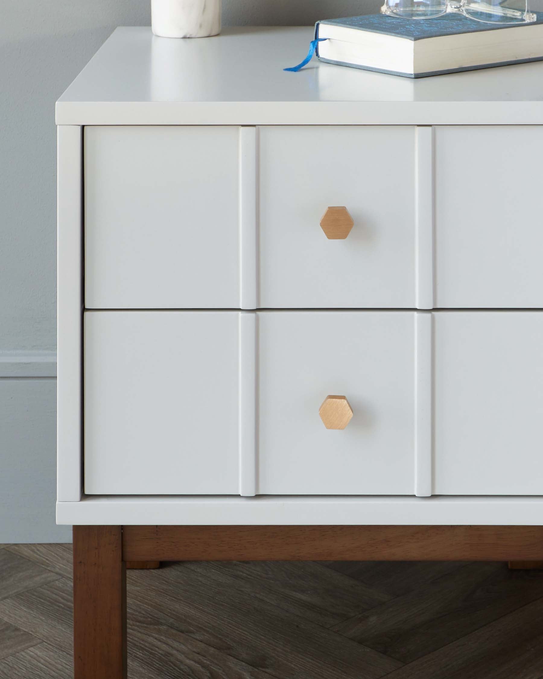 Modern white drawer chest with hexagonal wooden knobs and a minimalist design, standing on four wooden tapered legs in a room with herringbone parquet flooring.