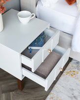 Contemporary white bedside table with two drawers, featuring sleek dark wood legs and stylish gold-tone drawer pulls. The top drawer is partially open, revealing a pair of eyeglasses and a book, while the bottom drawer displays neatly folded grey fabric. A white ceramic cup rests atop the table's smooth surface, beside a bed with white linens.