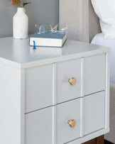 A modern white three-drawer bedside chest with wooden knobs. The top surface holds a white vase with a sprig of flowers, a closed book, and a pair of clear eyeglasses.