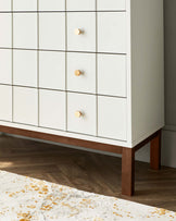 A modern white dresser with contrasting walnut legs and geometric wooden knobs. The dresser features multiple drawers with a subtle recessed grid pattern, providing a sleek and contemporary storage option. A partially visible textured rug with gold accents is in the foreground, indicating a stylish interior setting.