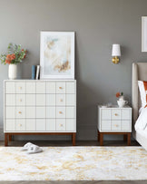 A modern bedroom furniture set featuring a large white dresser with multiple drawers accented by round wooden knobs and a smaller matching white nightstand with two drawers and wooden legs, both displayed on a textured cream and gold area rug.