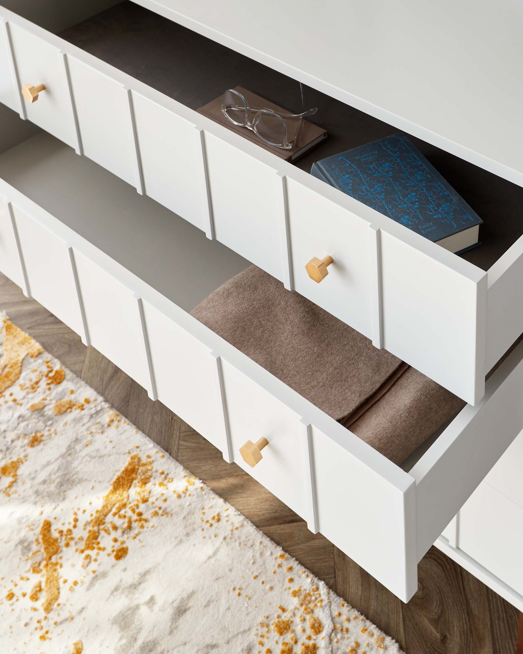 Modern minimalist white storage cabinet with multiple drawers, featuring sleek cut-out handles and light wooden pulls for a pop of natural contrast. Two of the drawers are partially open, displaying a neatly folded brown textile and a book with eyeglasses on top. The cabinet sits atop a hardwood floor with a corner of a textured rug showing earthy tones and an abstract pattern.