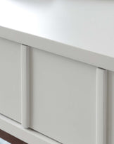 Close-up view of a modern, minimalist white drawer front with sleek, silver-tone handles against a white background.