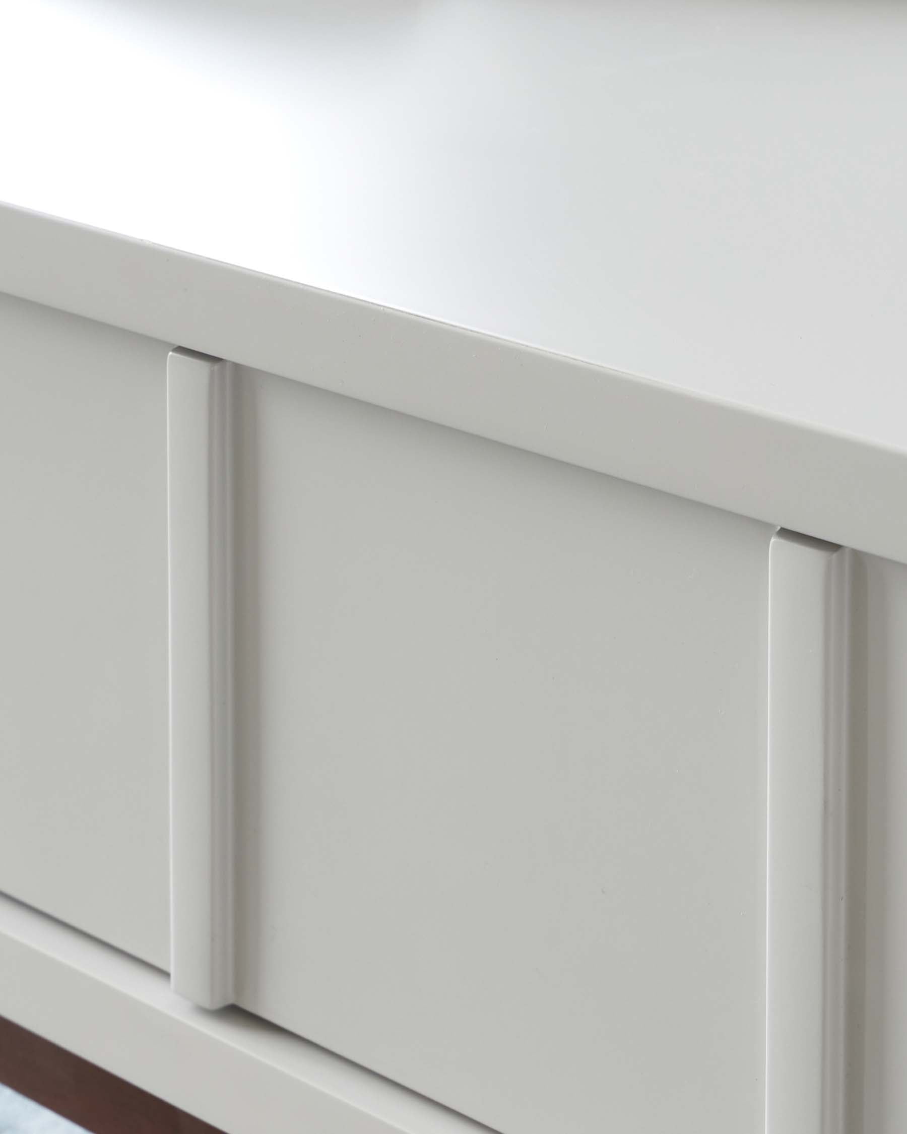 Close-up view of a modern, minimalist white drawer front with sleek, silver-tone handles against a white background.