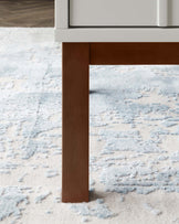 A close-up view of a modern furniture leg with a simple design, showcasing a white piece with a wooden, rectangular leg against a textured pale blue and white area rug, highlighting a contemporary and minimalist aesthetic.