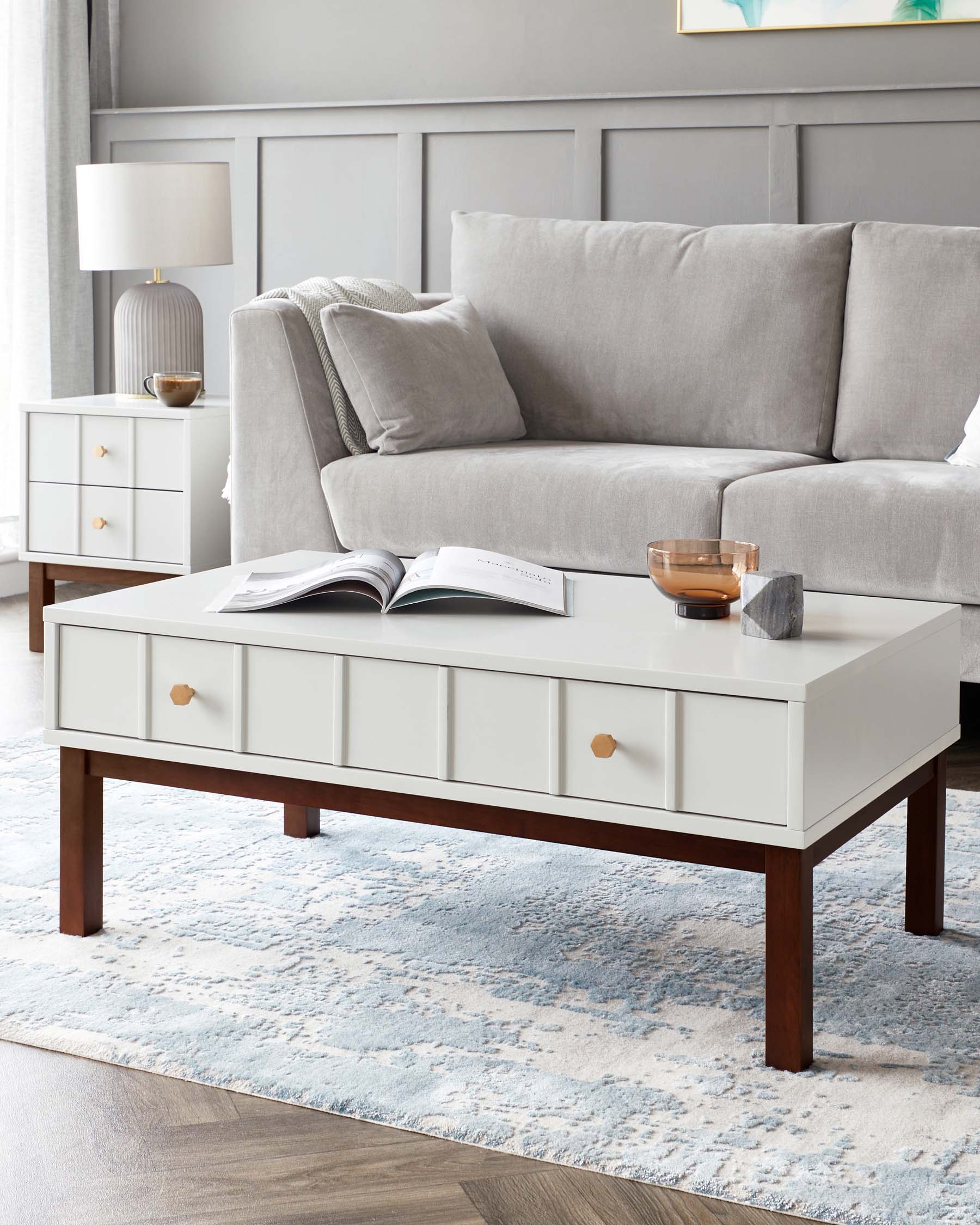 Elegant living room furniture set including a modern light grey upholstered sofa with plush cushions and a throw pillow, a white rectangular coffee table with four drawers featuring round brass knobs and dark brown tapering legs, and a matching white side table with two drawers and brass handles, all atop a textured blue and white area rug. A grey ceramic table lamp with a white shade complements the side table.