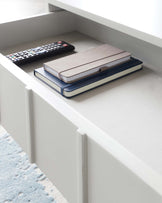 Modern white open shelf with clean lines, set against a light grey wall. The shelf is part of a larger unit and contains neatly stacked notebooks and a remote control, suggesting an organized living or office space.