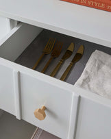 Modern white storage drawer featuring a sleek design with a brass hexagonal knob, partially open to reveal a neat arrangement of gold flatware and a linen napkin, set against a grey drawer mat.