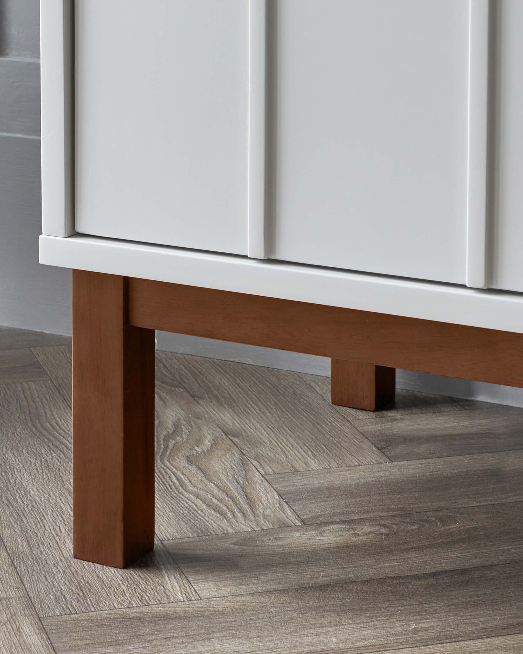 Close-up of a minimalist white cabinet with a clean design, featuring a modern, sleek white finish and sitting on sturdy, straight wooden legs in a rich brown tone, set against a hardwood floor with a grey woodgrain finish.