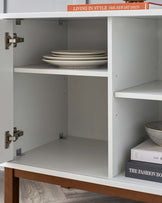 Minimalist white open shelving cabinet with visible hinges, featuring a single shelf partially occupied by a stack of dinner plates and another shelf with decorative books and a bowl, all displayed above a wooden base.