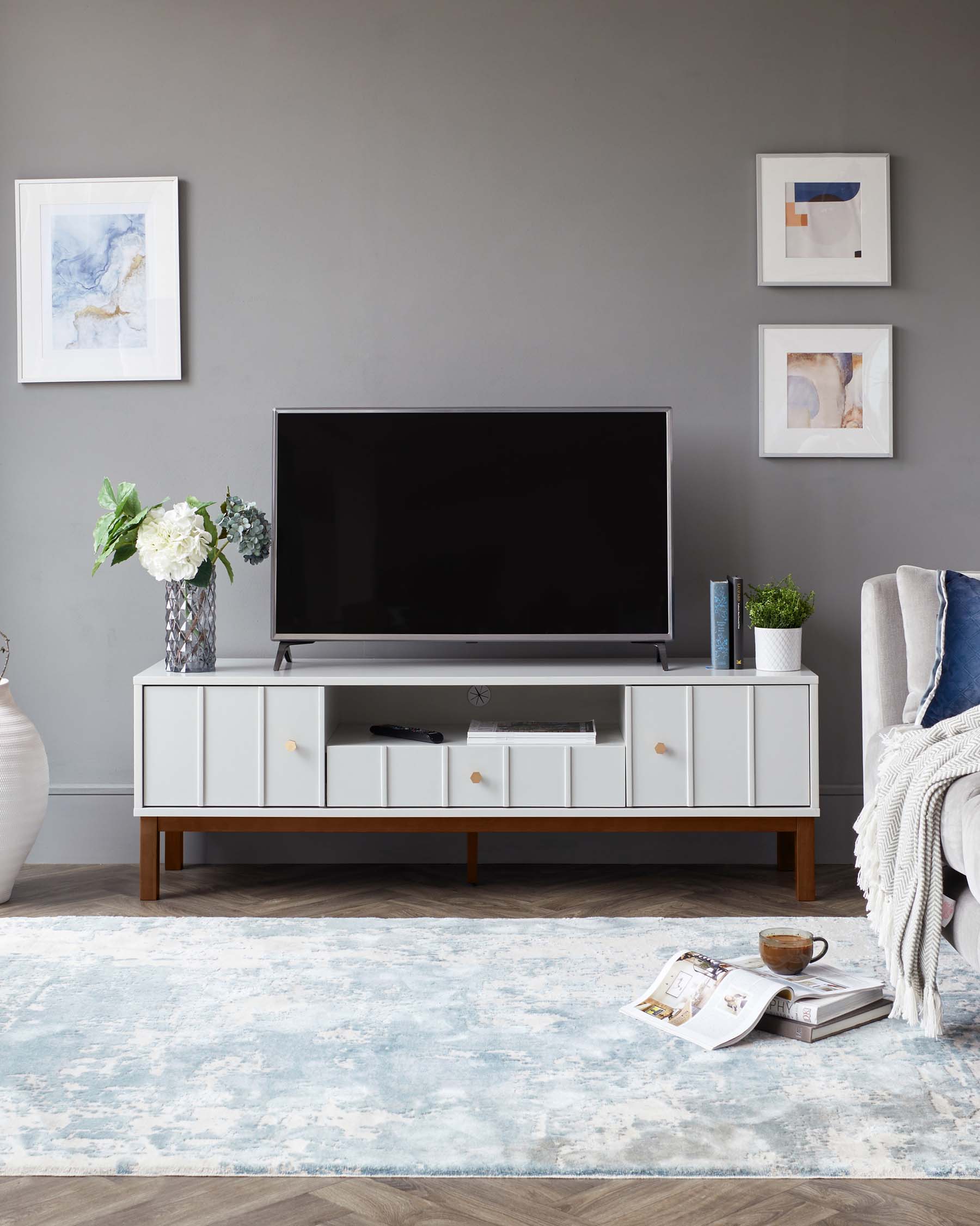 Mid-century modern white TV stand with tapered wooden legs and multiple storage compartments, including open shelves and closed drawers with gold-toned handles. A patterned blue and white area rug lies beneath it.