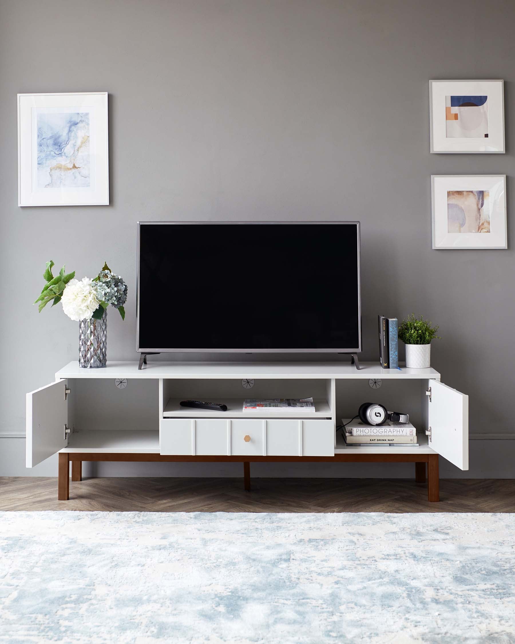 A modern white television stand with a spacious surface and multiple storage compartments, including open shelving and closed cabinets, supported by a sleek wooden base. The unit is accessorized with decorative items and placed on a patterned area rug. There is also a framed artwork above the TV stand on the wall.