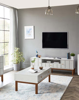 Modern furniture set in a living room, comprising of a white coffee table and a matching media console with a clean, minimalist design. Both pieces feature a combination of white surfaces with contrasting wooden legs. The coffee table is rectangle-shaped with drawers equipped with brass pull handles, while the media console also includes several drawers and open shelving, providing both storage and display options. The design reflects contemporary style with a nod to mid-century modern aesthetics.