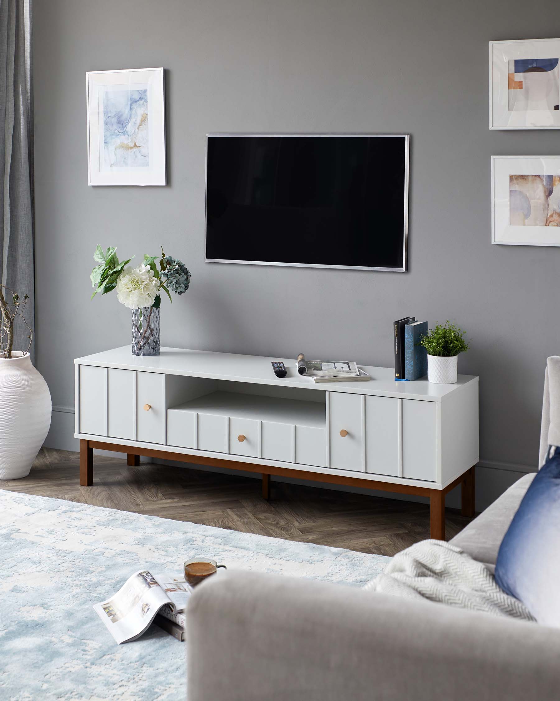 A contemporary white entertainment unit with a wooden base characterized by its sleek lines and minimalist design. The unit features two closed cabinet sections flanking a row of four drawers, all accented with round wooden knobs. A flat-screen television is mounted above the unit, which is decorated with a vase of white flowers and a small potted plant. A light grey fabric sofa is visible in the foreground, and the room is completed with a pale blue area rug on a wooden floor.