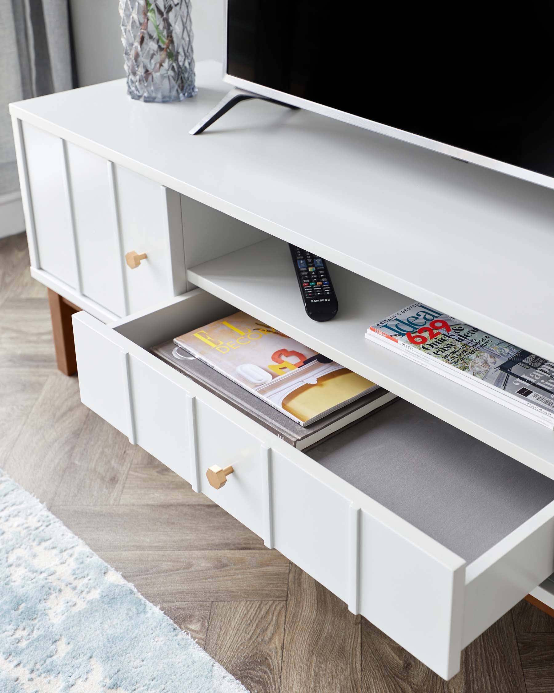 Modern white TV stand with spacious drawers and gold handles, featuring open shelving with magazines, remote control, and decorative vase atop a wood grain floor with a partial view of a light blue area rug.