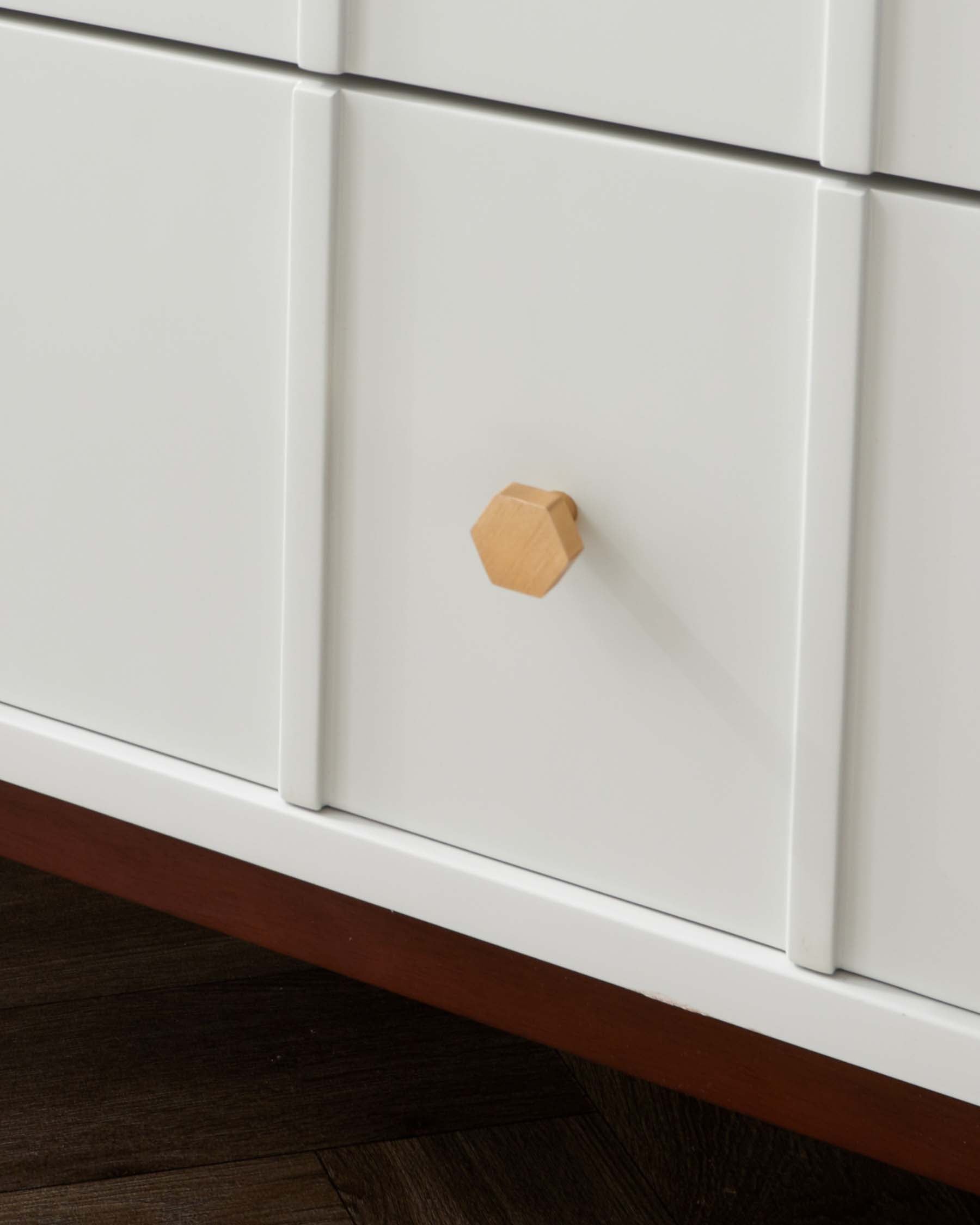 A close-up of a modern, white cabinet detail with a minimalist wooden knob and a dark wooden base against a hardwood floor.