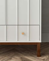 A modern white cabinet with a minimalist design, featuring clean lines, flat panel doors, and a single wooden knob on one of the doors. The piece stands on a short, flat wood base that provides a contrast to the all-white structure.