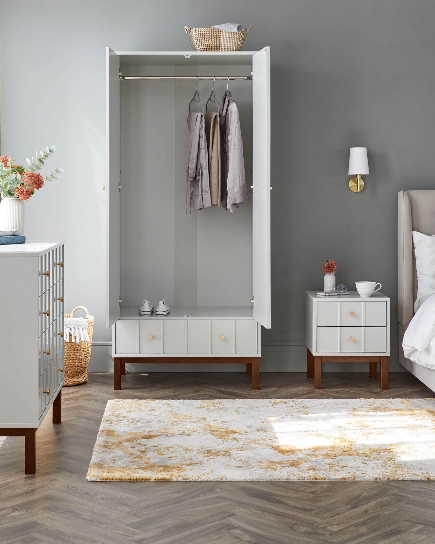 Contemporary matching bedroom furniture set against a grey wall, including a white wardrobe with an open door revealing a hanging rail with neatly placed clothes and a top shelf with a wicker basket. Beside it is a white bedside table with two drawers, wooden legs, and a lamp on top, complementing the nearby white sideboard with three drawers and two cabinets. The set is completed with chevron wood flooring and a white and gold patterned area rug.