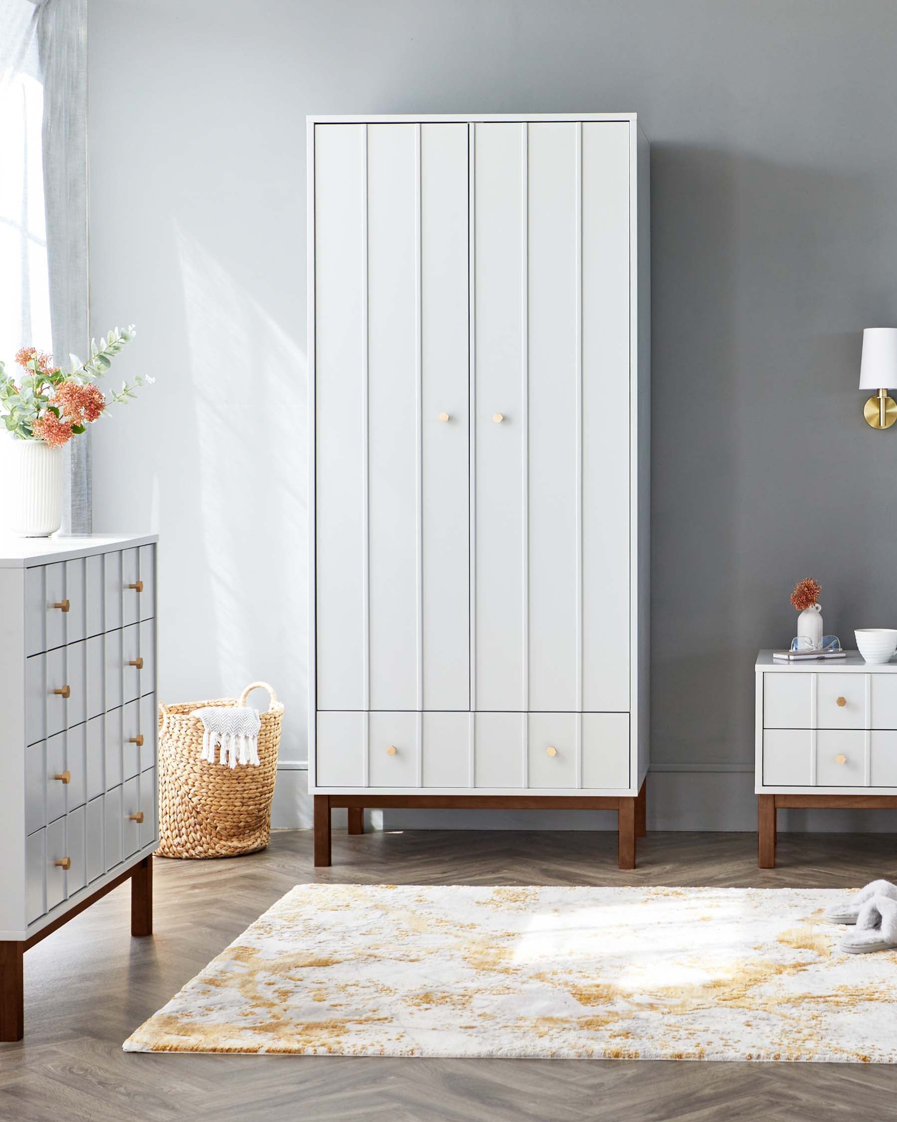 Elegant white bedroom furniture set including a tall wardrobe with golden knobs, a matching five-drawer dresser, and a two-drawer nightstand, all featuring clean lines and wooden legs, showcased in a room with soft grey walls and a plush white and gold area rug.