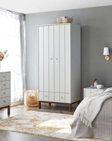 A tall white wardrobe with vertical panelling and golden knobs stands prominently against a grey wall. On the left, there's a white four-drawer chest with matching knobs and a wooden base. Both pieces have a minimalist style with a hint of mid-century modern influence due to the wooden accents. A woven basket rests atop the wardrobe, while another decorative basket sits on the floor beside the chest. A patterned area rug with white and gold hues stretches out in the foreground.