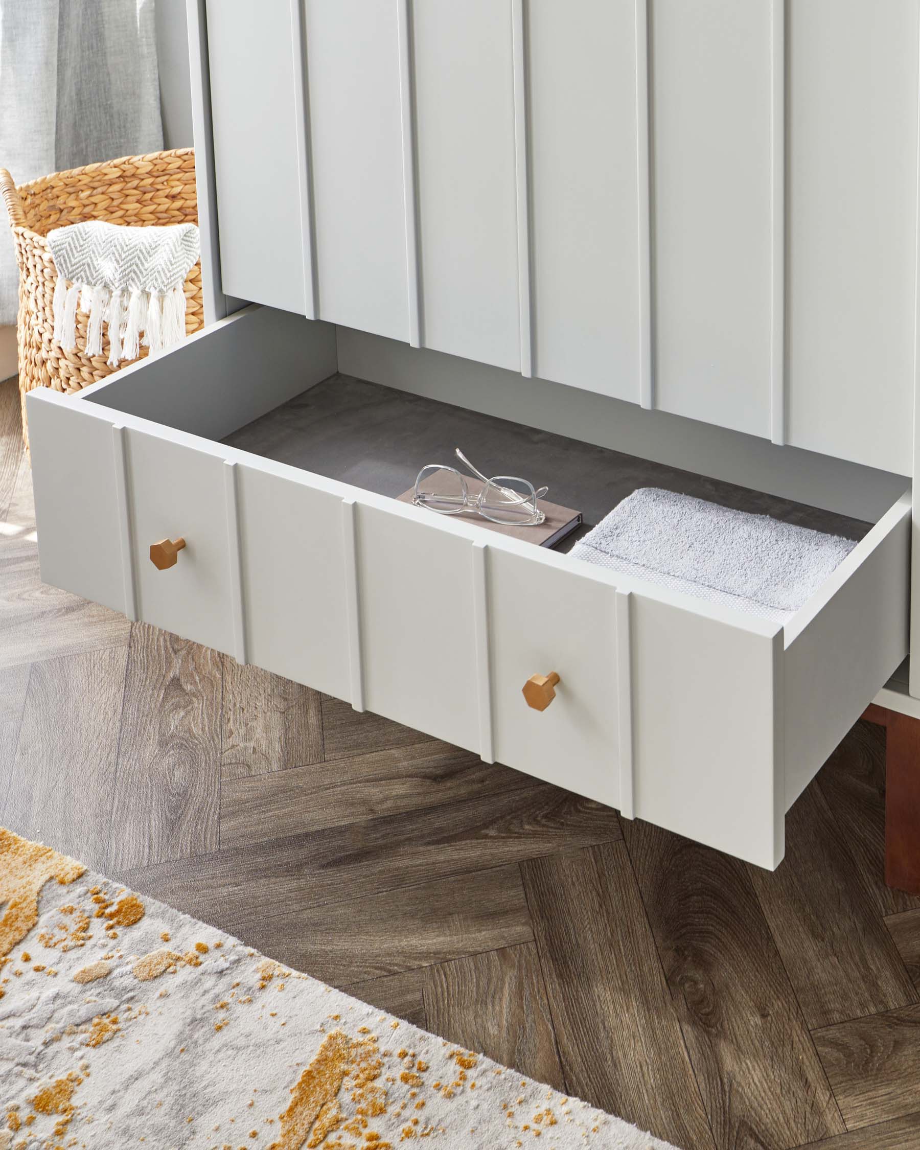 White modern storage bench with three drawers and leather pulls, featuring a cushioned seat, over a dark wood floor next to a wicker basket with a decorative throw.