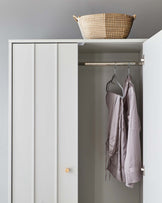 Minimalist white wardrobe with two-door front, a wooden knob on the right panel, and a metal clothes rail inside holding a single coat. A wicker basket sits atop the wardrobe against a grey wall.