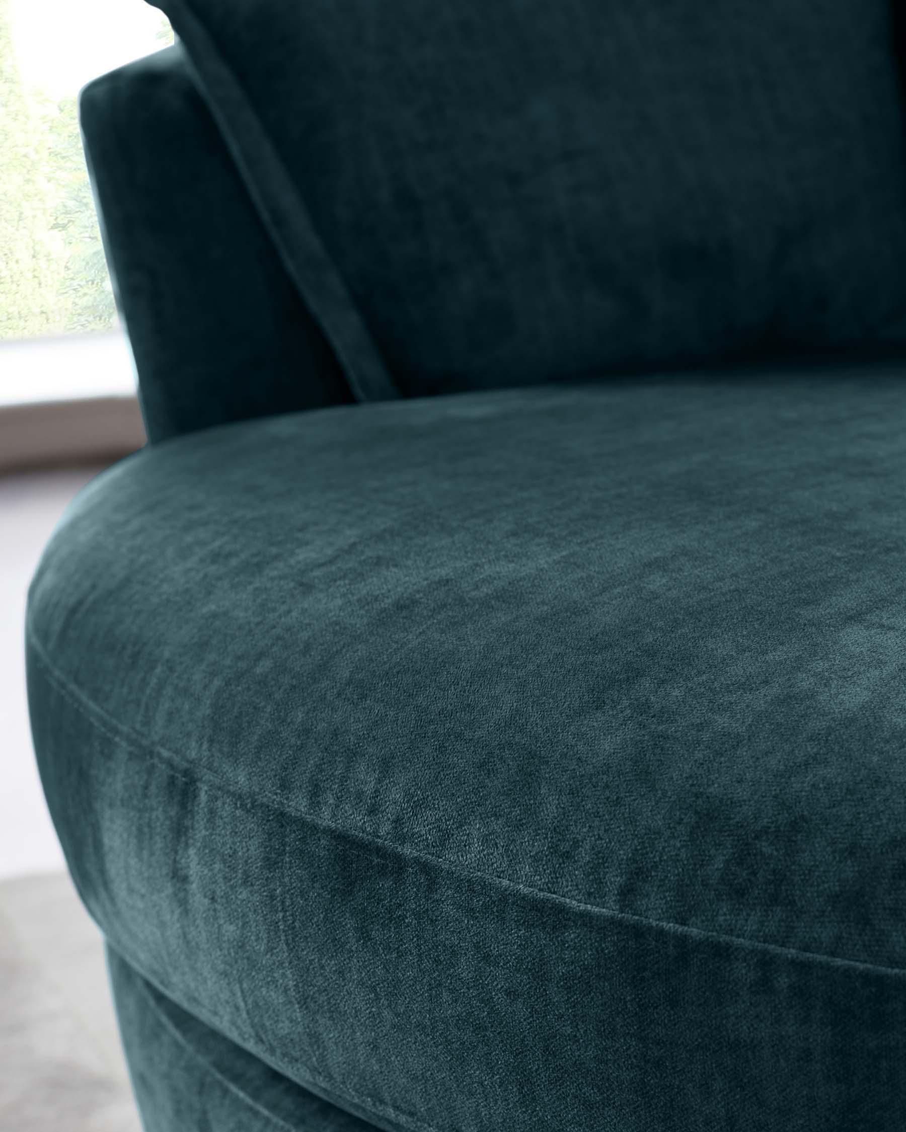 Close-up of a luxurious navy blue velvet armchair with plush upholstery and a smooth, rounded profile, positioned against a light-filled window with a greenery view.