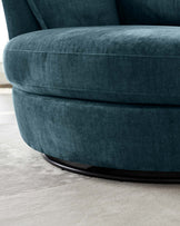 Plush deep teal upholstered armchair with a smooth, curved design and a visible seam detail running along the seat cushion. The chair rests on a low-profile, circular black base.