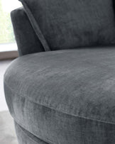 Close-up of a contemporary dark grey upholstered armchair with a plush seat cushion and soft fabric texture, partially visible armrest, with a blurred natural light background indicating a window.